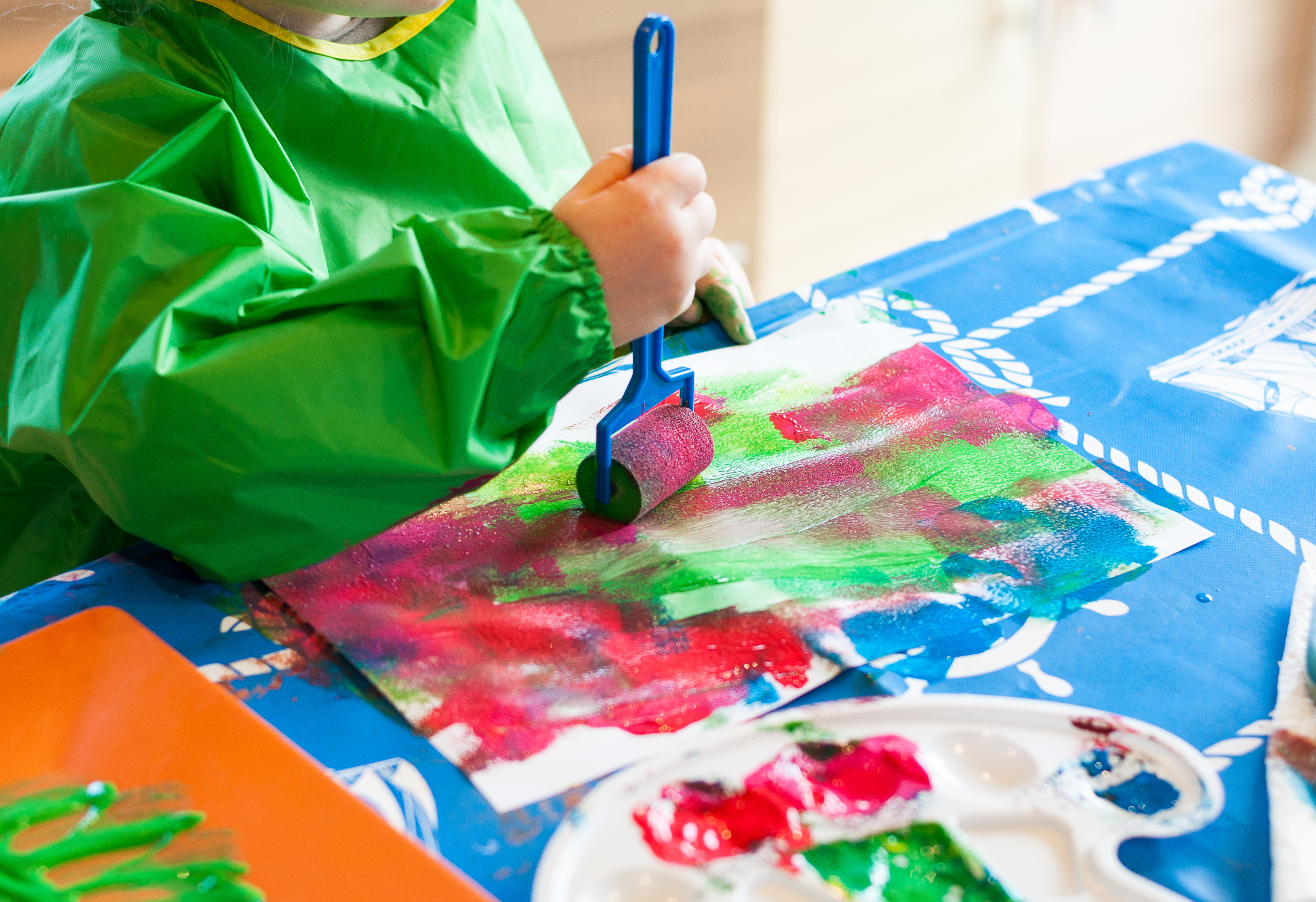 Photo of a child in a green apron painting red and green on a piece of paper with a roller
