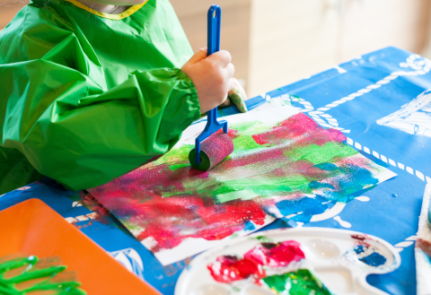 Photo of a child in a green apron painting red and green on a piece of paper with a roller